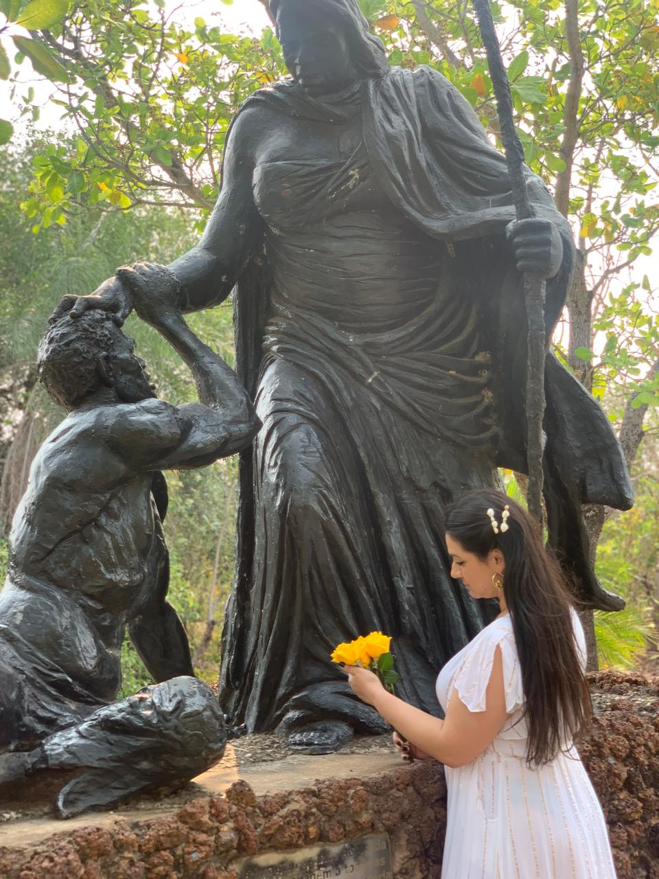 Caminhos de Luz vestida de branco, com flores nas mãos, diante de uma imagem sagrada na mata